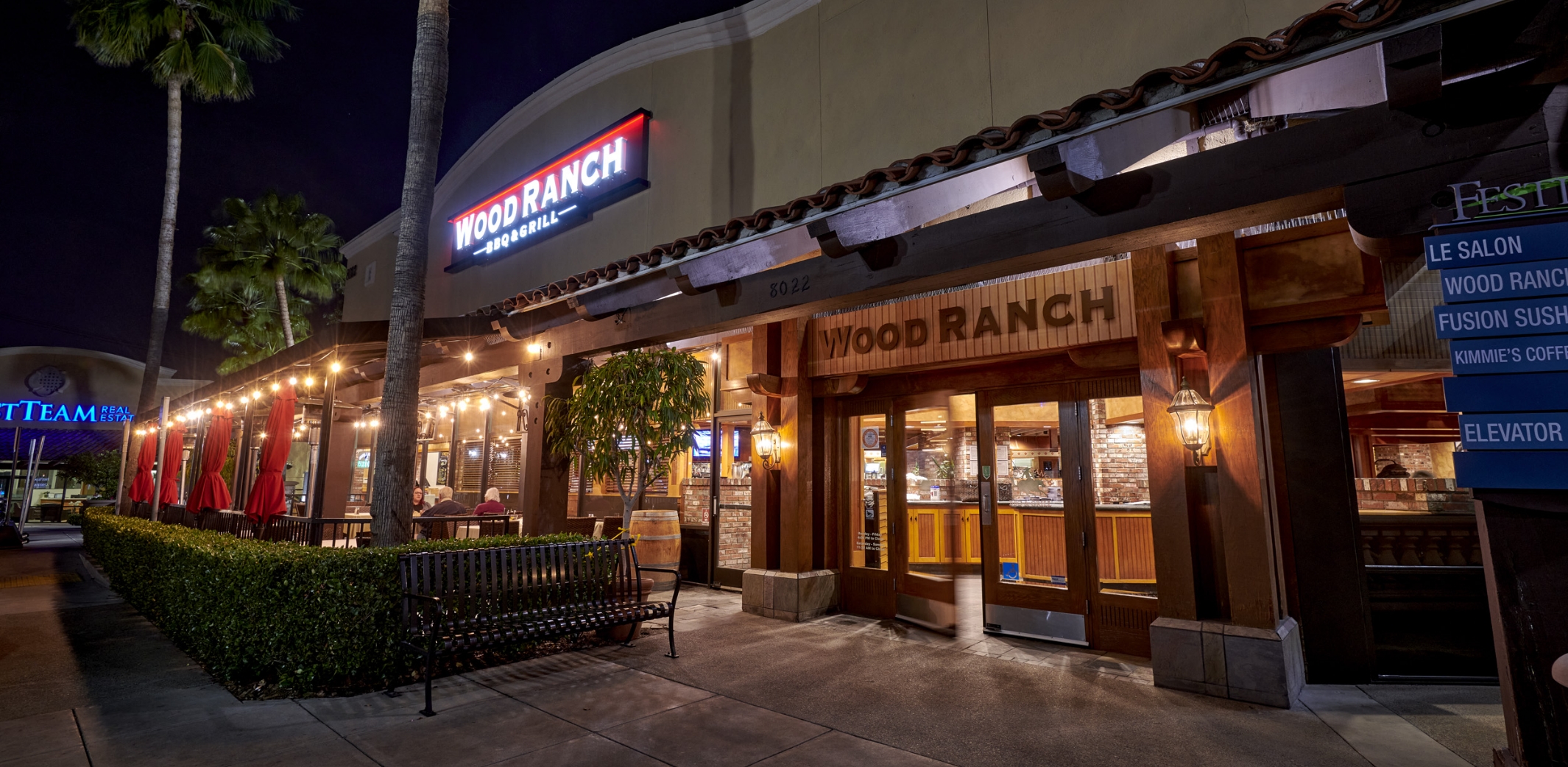 Exterior view of Wood Ranch BBQ & Grill Aneheim in the evening, featuring warm lighting and a welcoming entrance with lush palm trees and an inviting outdoor seating area