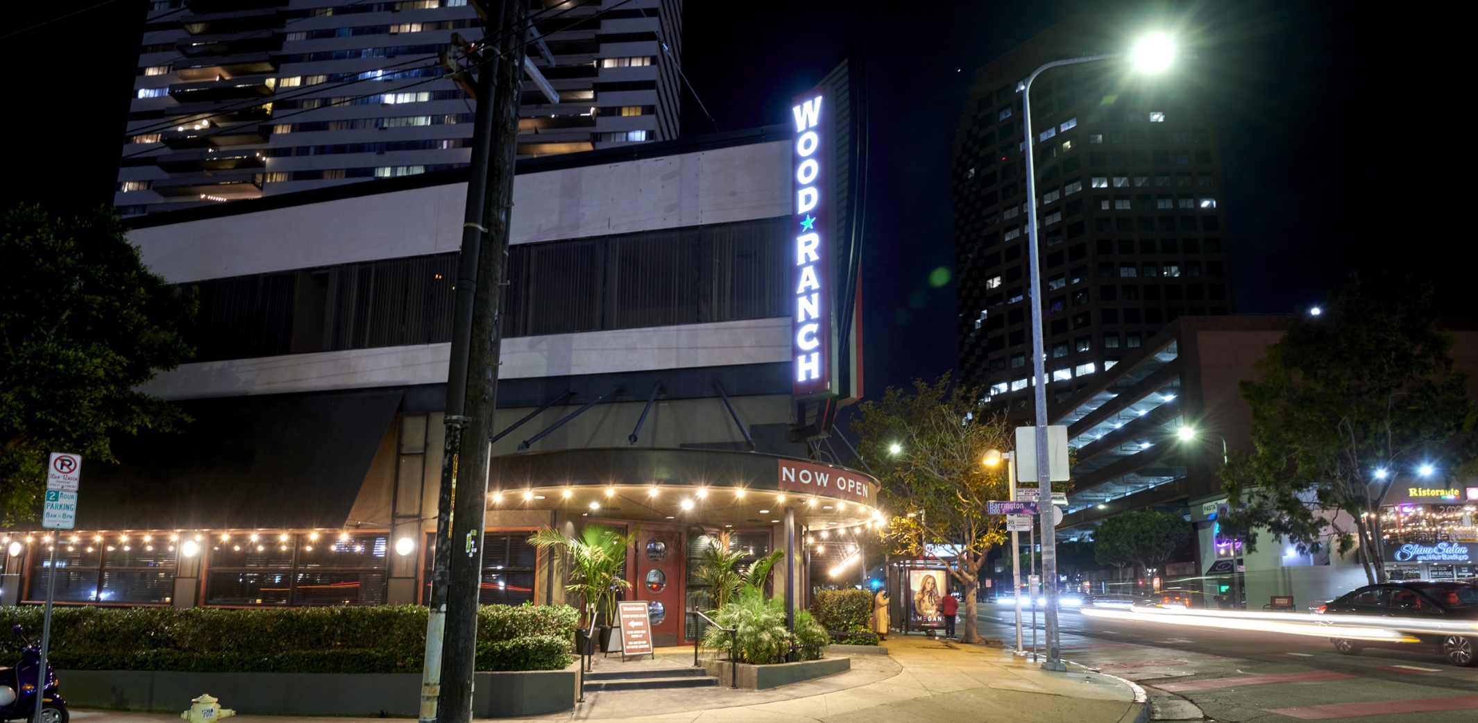 Nighttime exterior of Wood Ranch BBQ & Grill, with its bright neon sign, illuminated patio area, and modern urban surroundings.