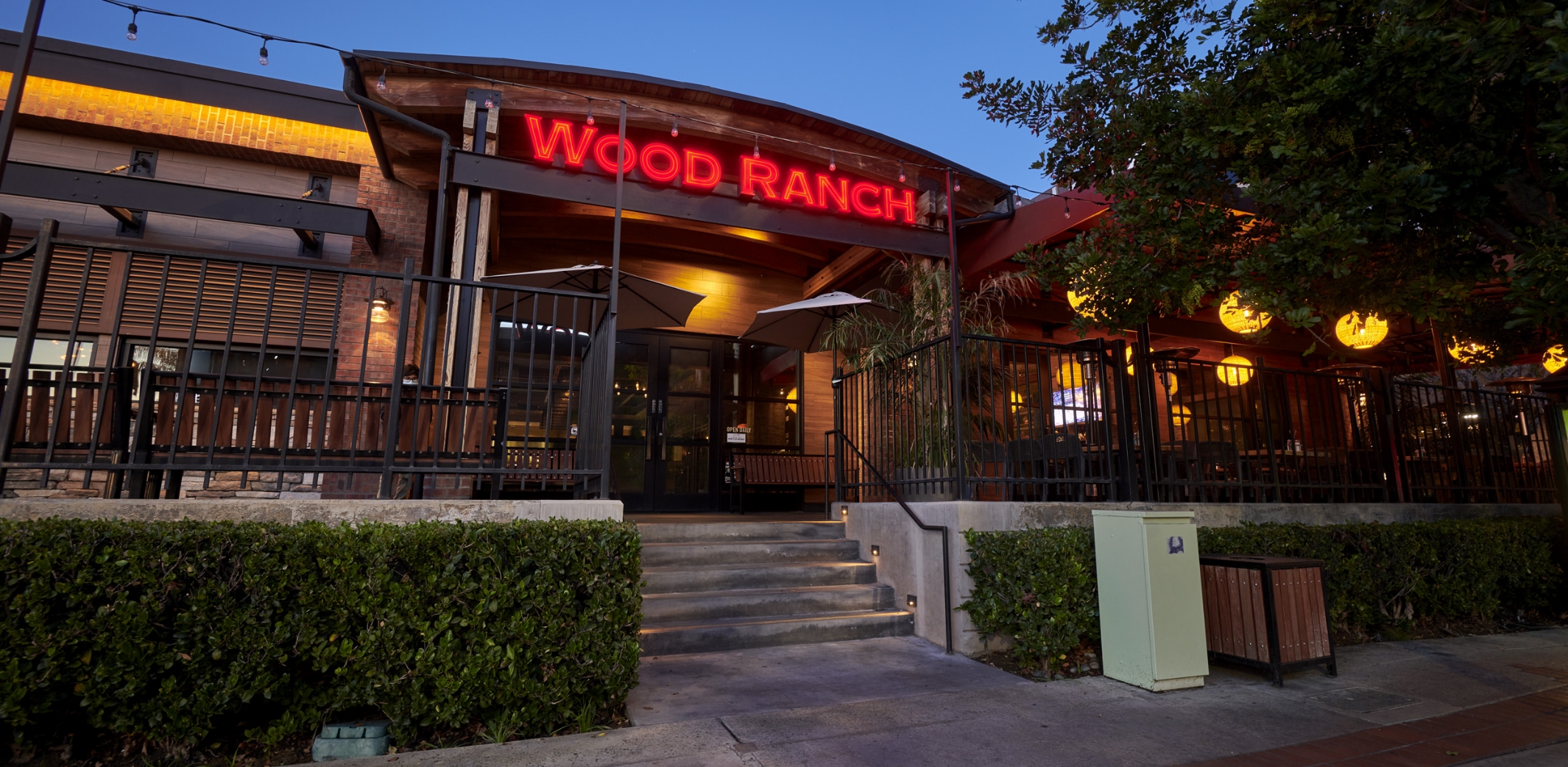 The exterior of Wood Ranch BBQ & Grill Burbank at dusk, showcasing the restaurant's illuminated sign and outdoor patio area with a cozy, welcoming ambiance.
