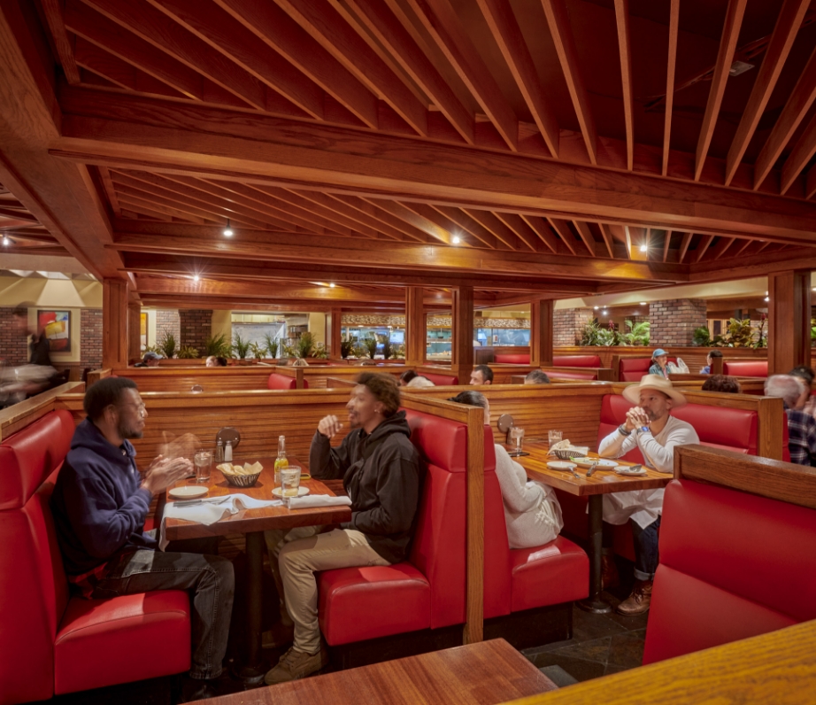 Diners seated in comfortable red booths under an intricate ceiling of wooden beams, enjoying meals and conversation in a warmly lit restaurant.