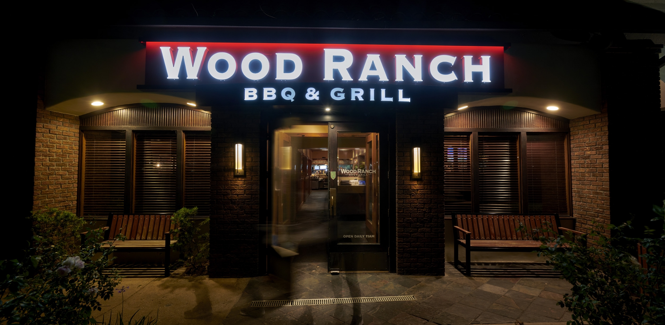Night view of Wood Ranch BBQ & Grill' Camarillo's inviting entrance, with its neon sign, warm interior lighting, and lush plants framing the doorway.