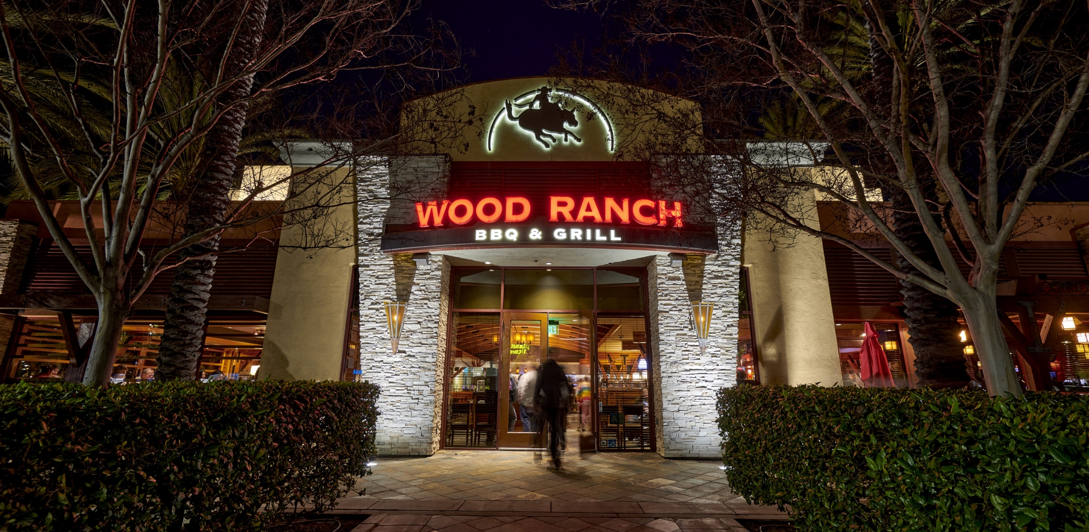 Exterior view of Wood Ranch BBQ & Grill at night with illuminated signage and a warmly lit entrance. Trees and neatly trimmed hedges frame the building, creating a welcoming atmosphere for diners.