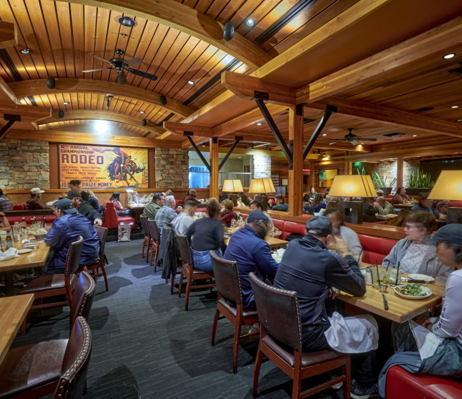 Interior of Wood Ranch BBQ & Grill showcasing a lively dining area with patrons seated at booths under a ceiling with warm wooden beams and ambient lighting.