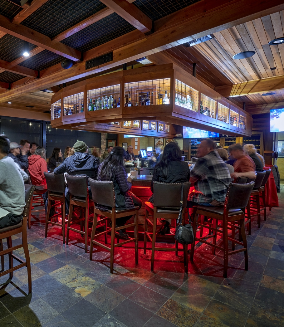 Patrons enjoying drinks and conversation at the bar in Wood Ranch BBQ & Grill, which features a central bar area with high stools, surrounded by multiple TV screens and a display of assorted beverages.