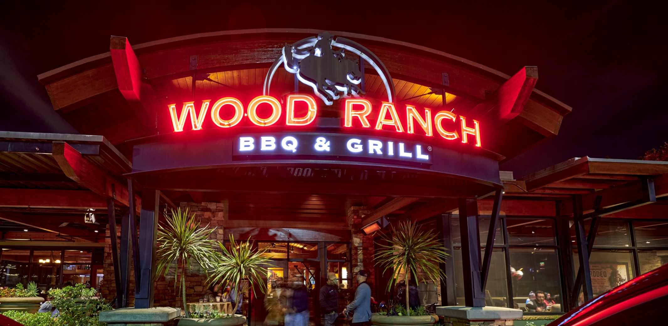 Nighttime view of the entrance to Wood Ranch BBQ & Grill in Chino Hills, with vibrant neon signage and guests entering, highlighting the restaurant's busy and welcoming environment.