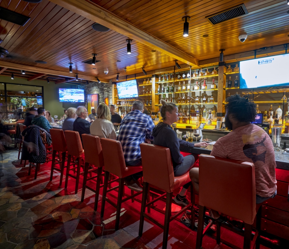 Interior view of Wood Ranch BBQ & Grill with patrons seated at red bar stools, enjoying the lively ambiance and watching televised sports above the bar.