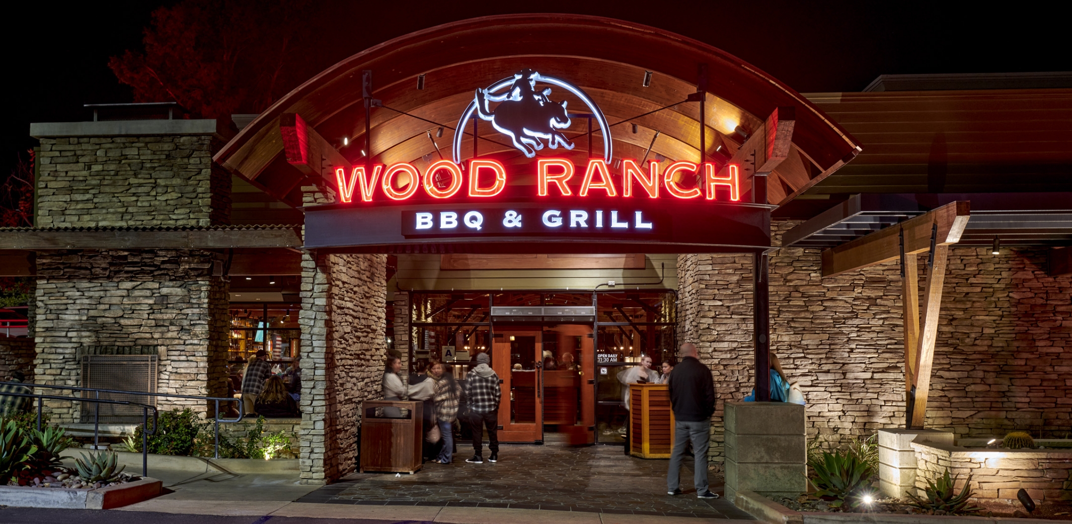 The inviting entrance of Wood Ranch BBQ & Grill Corona at night, featuring the restaurant's radiant red neon sign and bustling activity as guests arrive.