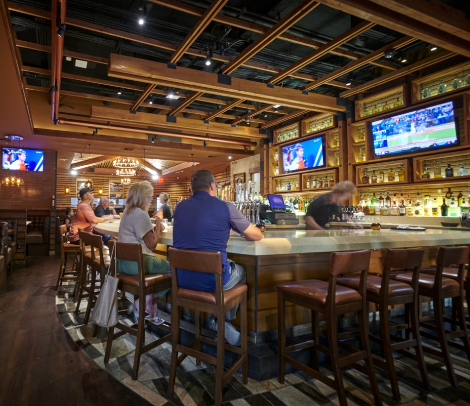 Patrons seated at the bar inside Wood Ranch BBQ & Grill, enjoying the lively atmosphere with multiple sports broadcasts on the overhead televisions, complemented by the warm wood finish of the bar area.