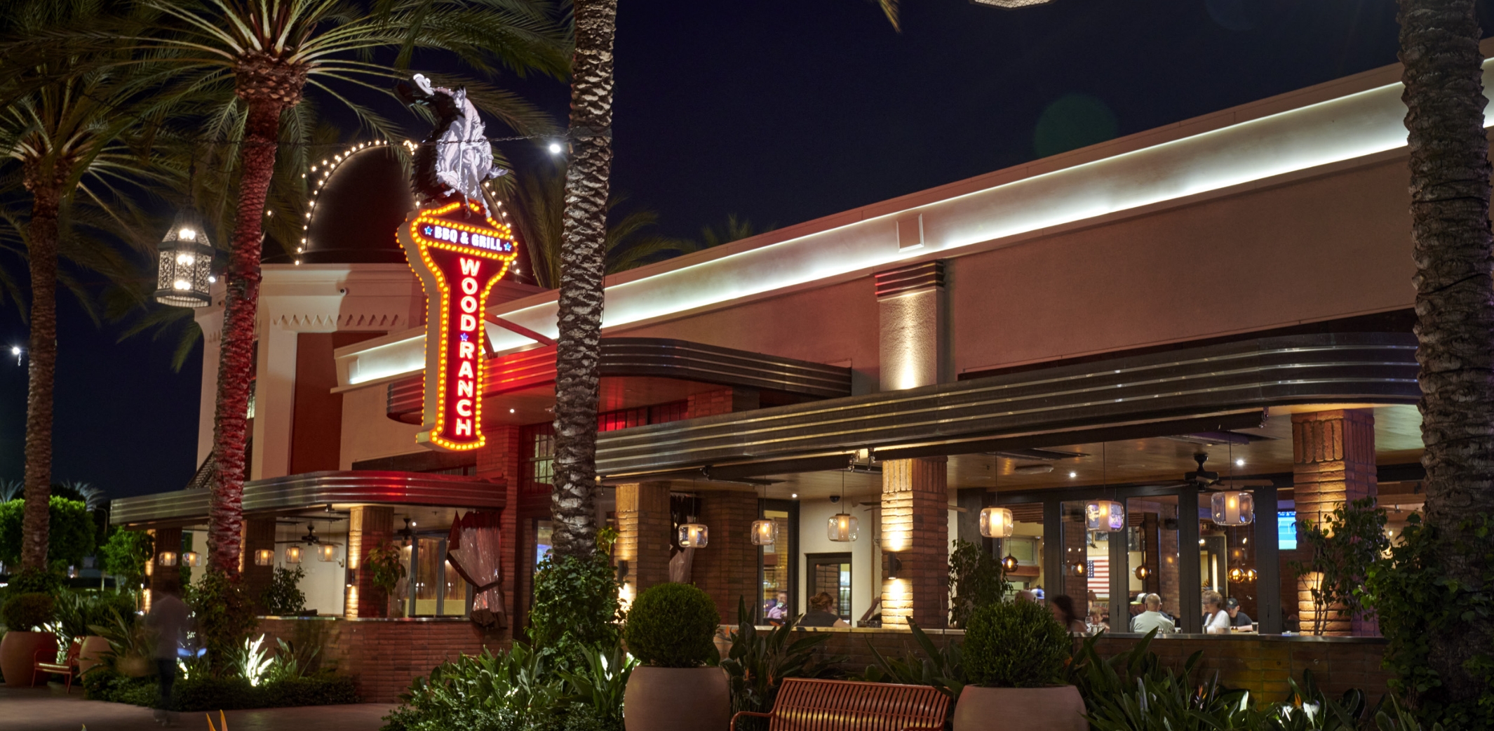 The iconic entrance of Wood Ranch BBQ & Grill Irvine at dusk, featuring neon signage and statuesque palm trees, inviting diners into the warmly lit, welcoming space.