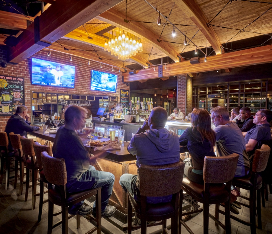 Patrons enjoy the ambiance of Wood Ranch BBQ & Grill's bar area with multiple TV screens displaying sports, wooden beams overhead, and a bustling atmosphere.