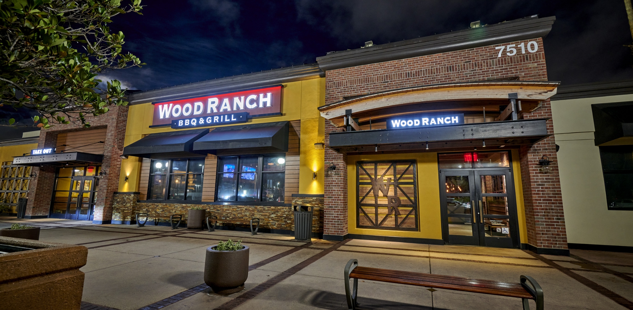 Exterior view of Wood Ranch BBQ & Grill in Mission Valley at night, showcasing the restaurant's bold signage and inviting entrance with a contemporary architectural design.