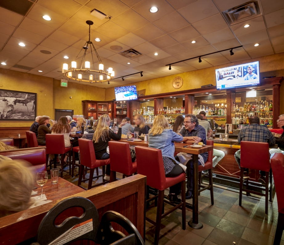 A bustling bar scene at Wood Ranch BBQ & Grill with patrons engaged in conversation, enjoying drinks and sports playing on TVs overhead, creating a social and inviting atmosphere.