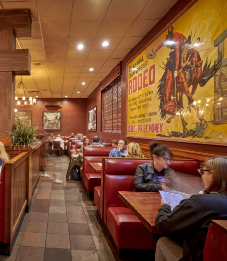 Diners seated in red booths at Wood Ranch BBQ & Grill, enjoying their meals beside a large, vibrant rodeo-themed mural, contributing to the restaurant's Western ambiance.