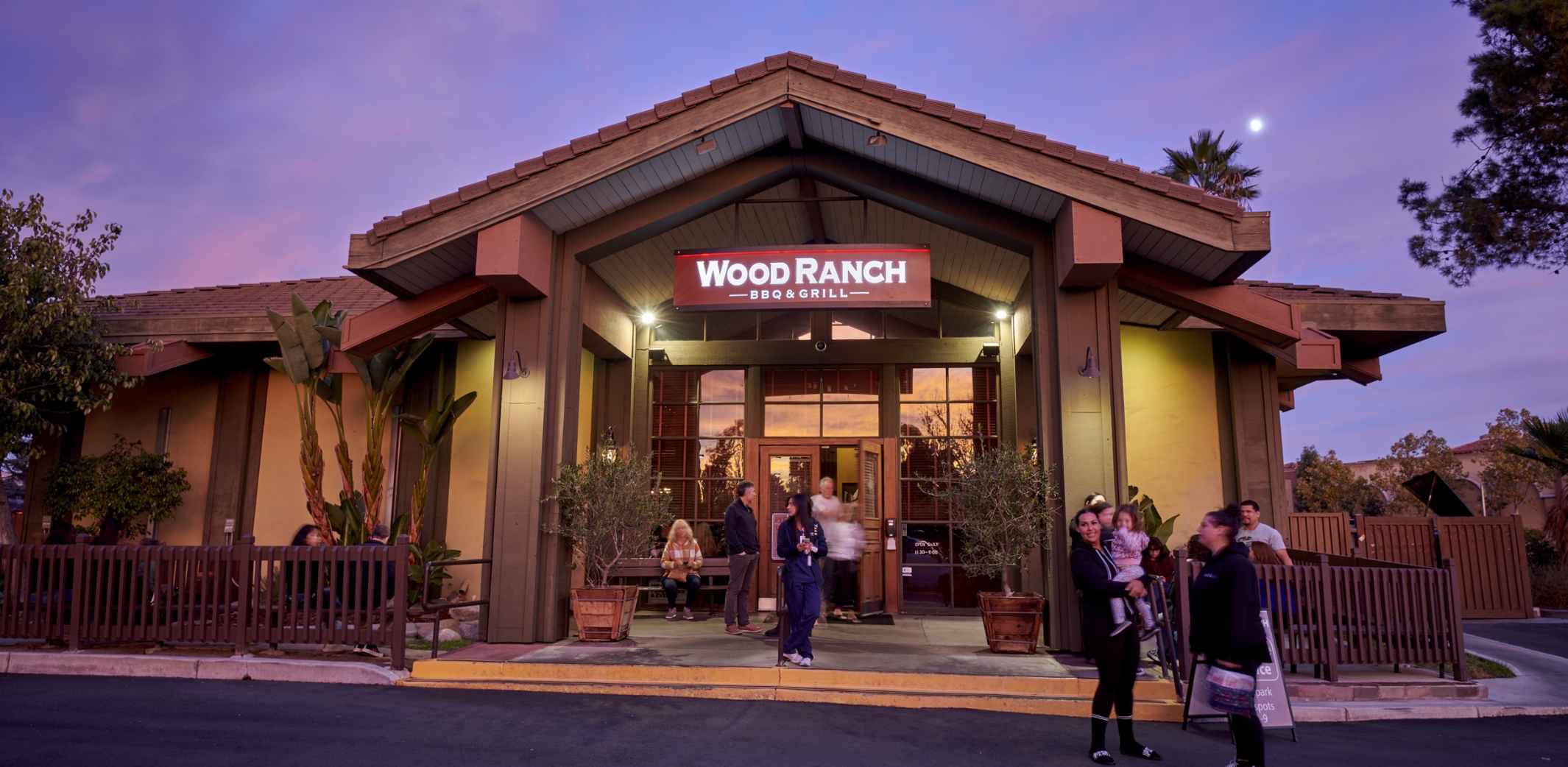 Twilight view of the Wood Ranch BBQ & Grill Moorpark entrance, with guests milling about, highlighted by the restaurant's warm lighting and welcoming signage.