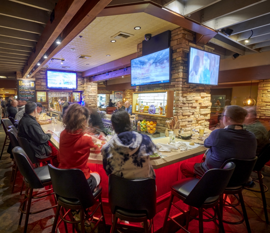 Patrons seated at a bar counter in Wood Ranch BBQ & Grill, engaging with each other with a backdrop of TV screens and a well-lit bar showcasing an array of beverages.