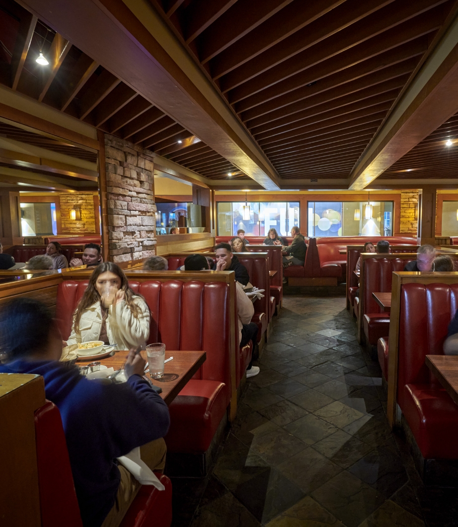 Customers enjoying their dining experience at Wood Ranch BBQ & Grill, comfortably seated in red booths within a spacious interior accented by wood finishes and ambient lighting.