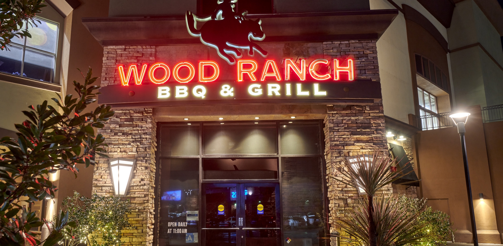 The front view of Wood Ranch BBQ & Grill at Northridge during evening hours, featuring the iconic neon-lit sign and modern architecture, with guests entering and exiting the restaurant.