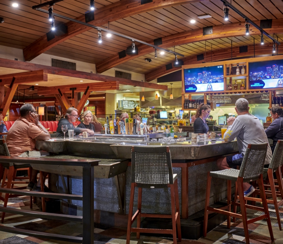 Customers gathered at the bar in Wood Ranch BBQ & Grill, enjoying a casual dining experience with sports games on the television screens in a warmly lit wooden interior.