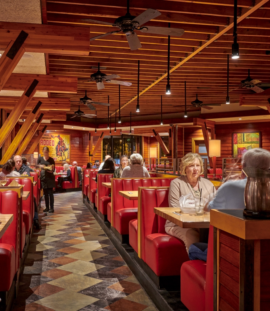 Patrons dining in red leather booths at Wood Ranch BBQ & Grill, with the restaurant's cozy wooden architecture and ambient lighting enhancing the comfortable atmosphere.