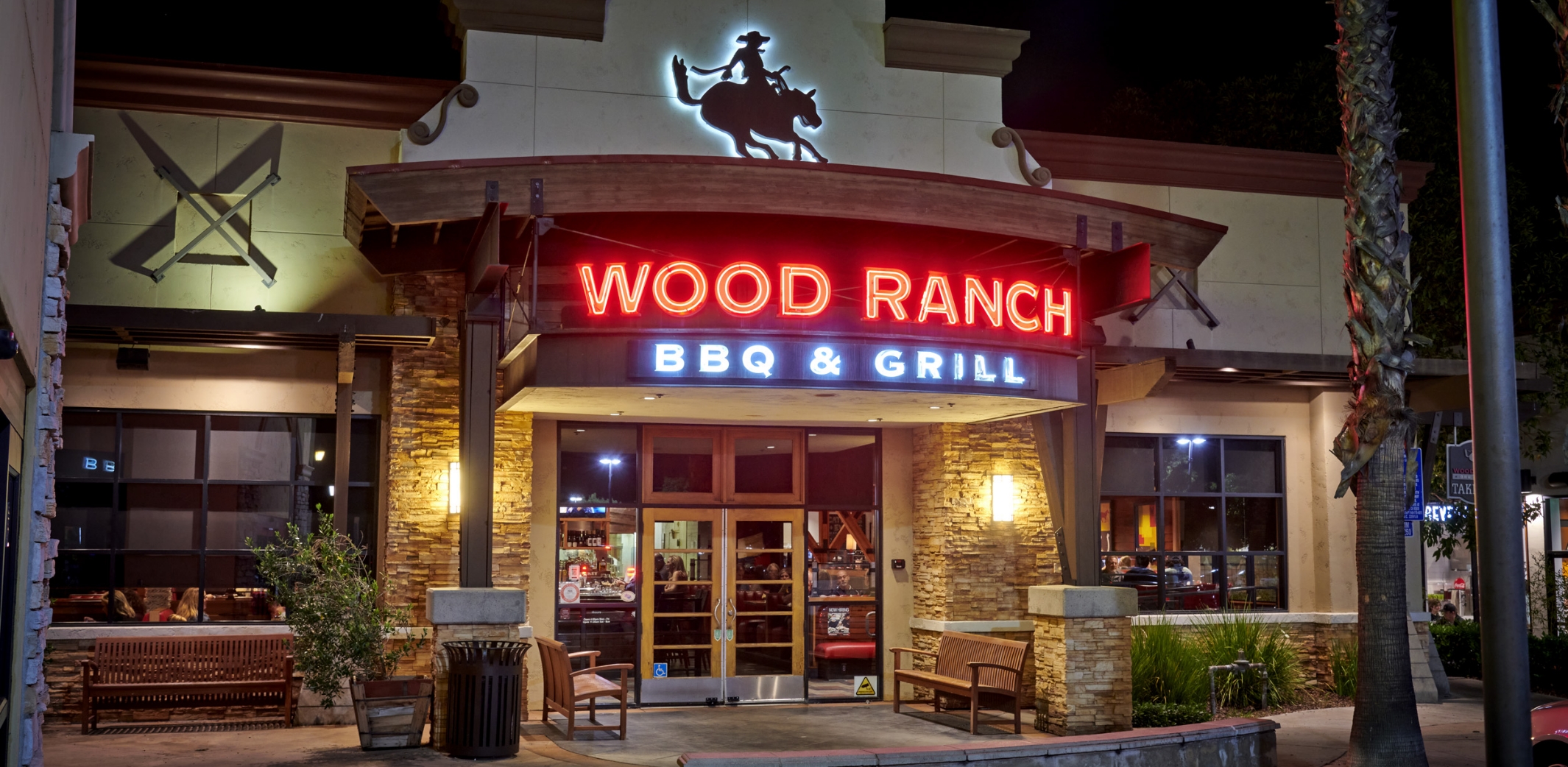 The inviting entrance of Wood Ranch BBQ & Grill Santa Margarita at night, featuring neon signage and a rustic stone facade, with guests arriving and departing under the soft glow of exterior lights.
