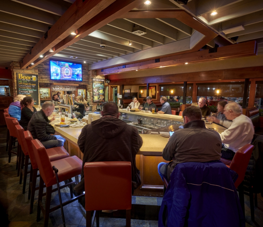 Guests seated around a central bar at Wood Ranch BBQ & Grill in Valencia, with sports broadcasts on TVs and a warm, inviting atmosphere highlighted by the wooden ceiling and ambient lighting.
