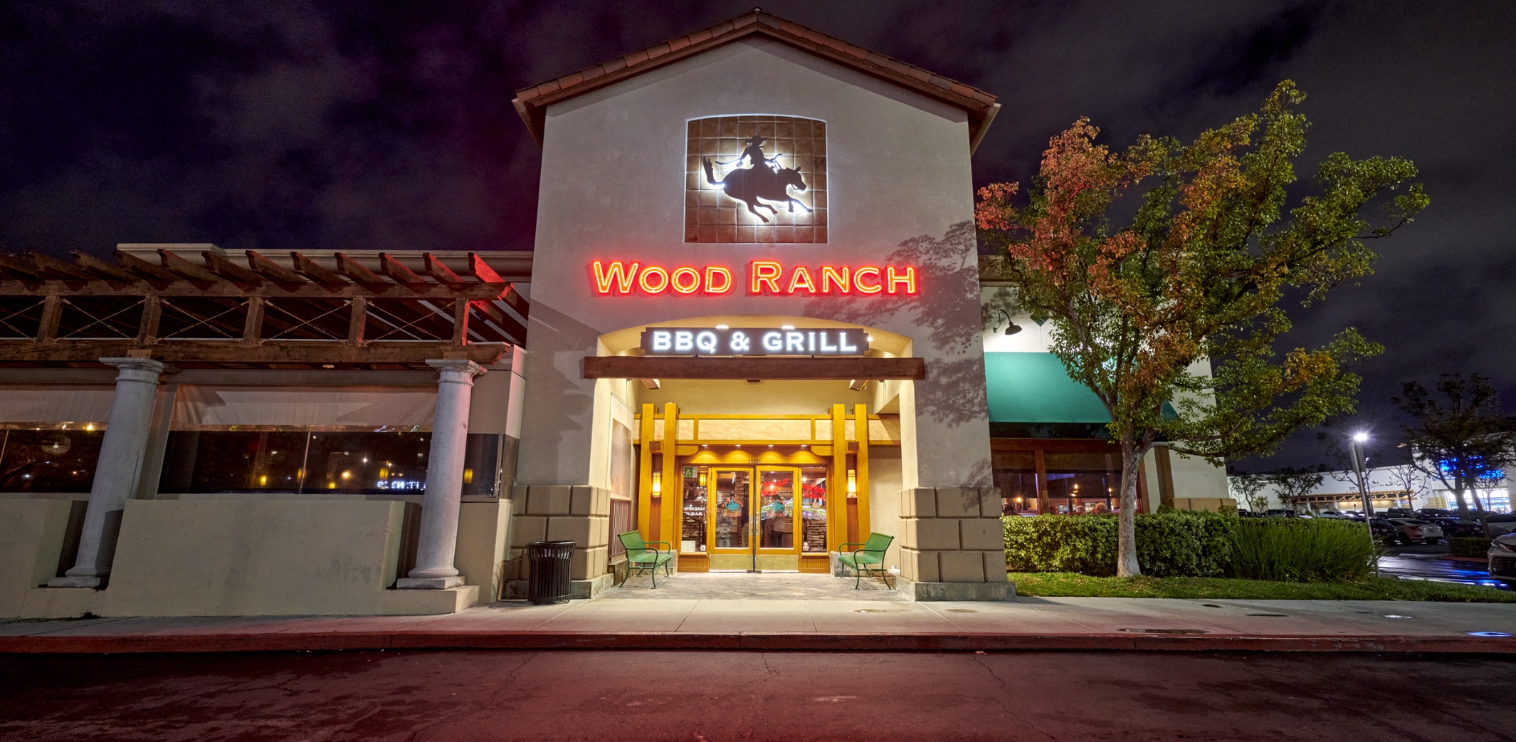 The illuminated facade of Wood Ranch BBQ & Grill in Valencia at night, showcasing its striking sign and welcoming entrance framed by mature trees and soft exterior lighting.