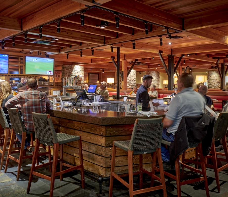 Patrons seated at the bar in Wood Ranch BBQ & Grill, Ventura, enjoying a convivial atmosphere with overhead sports telecasts and a rustic, wood-dominated décor.