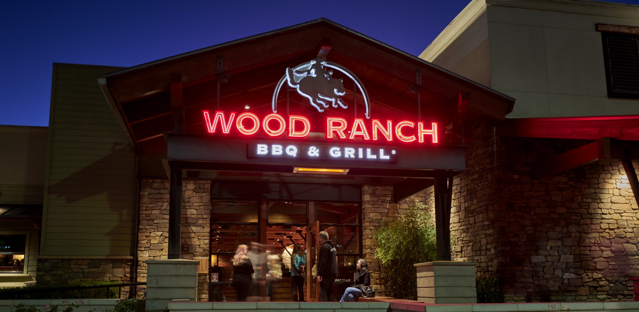 The exterior of Wood Ranch BBQ & Grill in Ventura at twilight with the restaurant's neon sign glowing, welcoming guests into the cozy, stone-clad building.