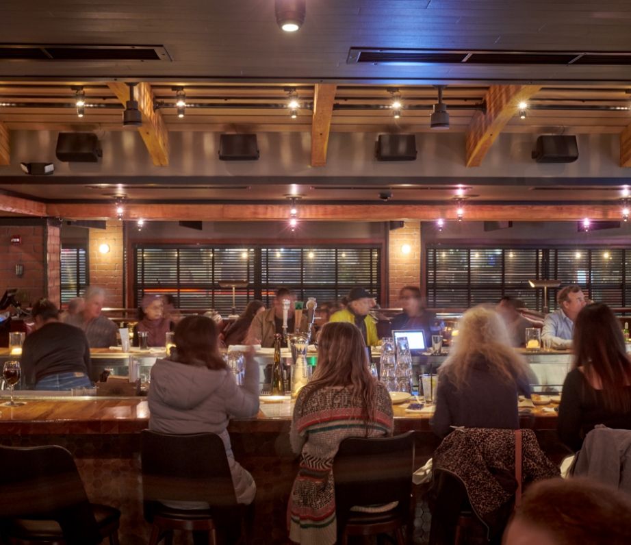 The interior of a restaurant with a vibrant bar scene, guests seated on stools, enjoying drinks and casual conversation under warm, ambient lighting.