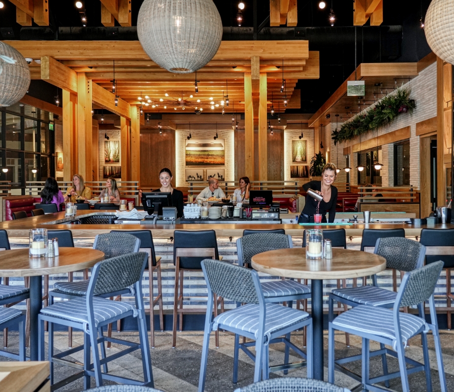 Interior view of Wood Ranch BBQ & Grill in Thousand Oaks with patrons seated at modern tables, a spacious bar area in the background, under elegant orb lighting and a high ceiling with wooden beams.