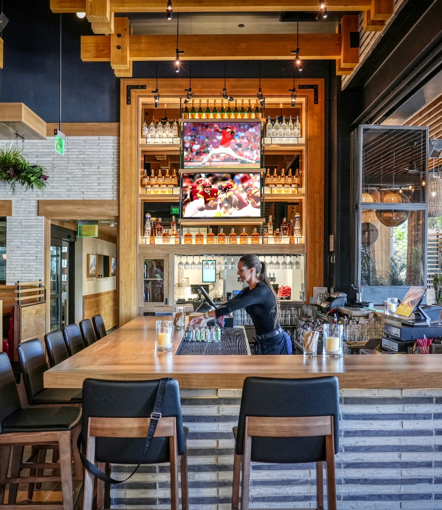 A bartender at Wood Ranch BBQ & Grill in Thousand Oaks prepares drinks at a sleek bar backed by a wall-mounted liquor display and multiple TV screens, amidst a setting of chic decor and ambient lighting.