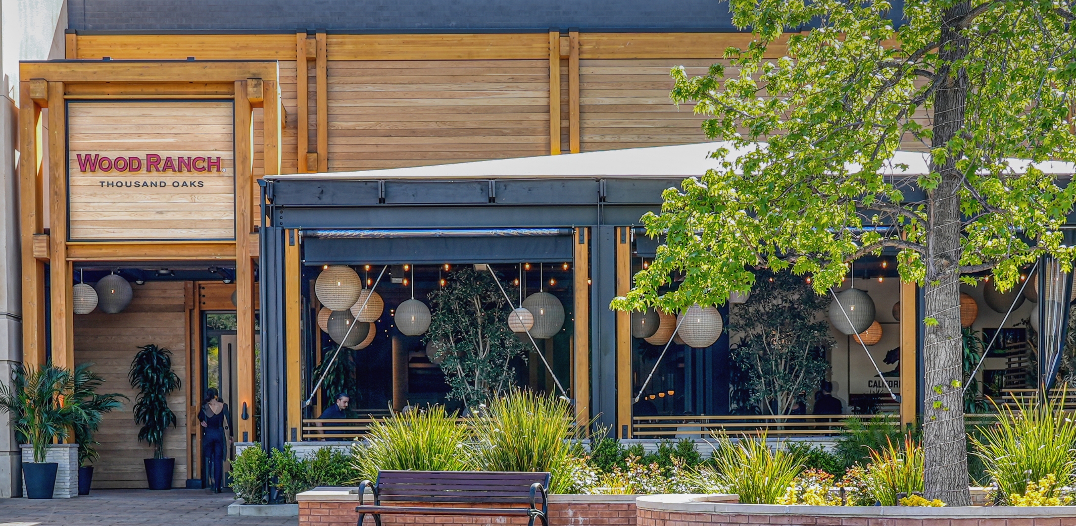 The front view of Wood Ranch BBQ & Grill in Thousand Oaks, displaying its name against a backdrop of contemporary architecture with large windows, and surrounded by vibrant greenery and outdoor seating.