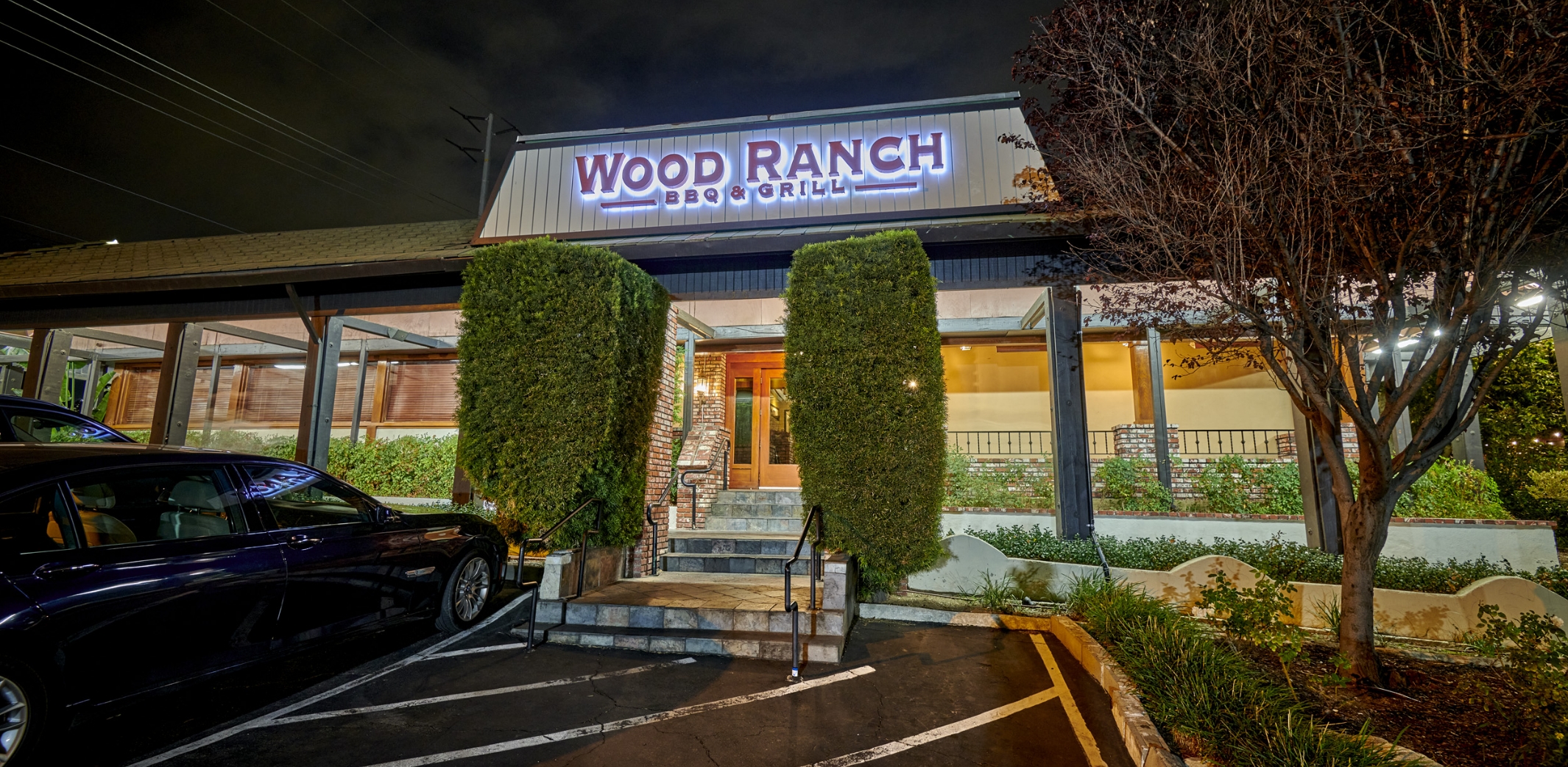 An evening view of Wood Ranch BBQ & Grill's exterior, featuring illuminated signage, neatly trimmed hedges, a brick stairway entrance, and a parked car.