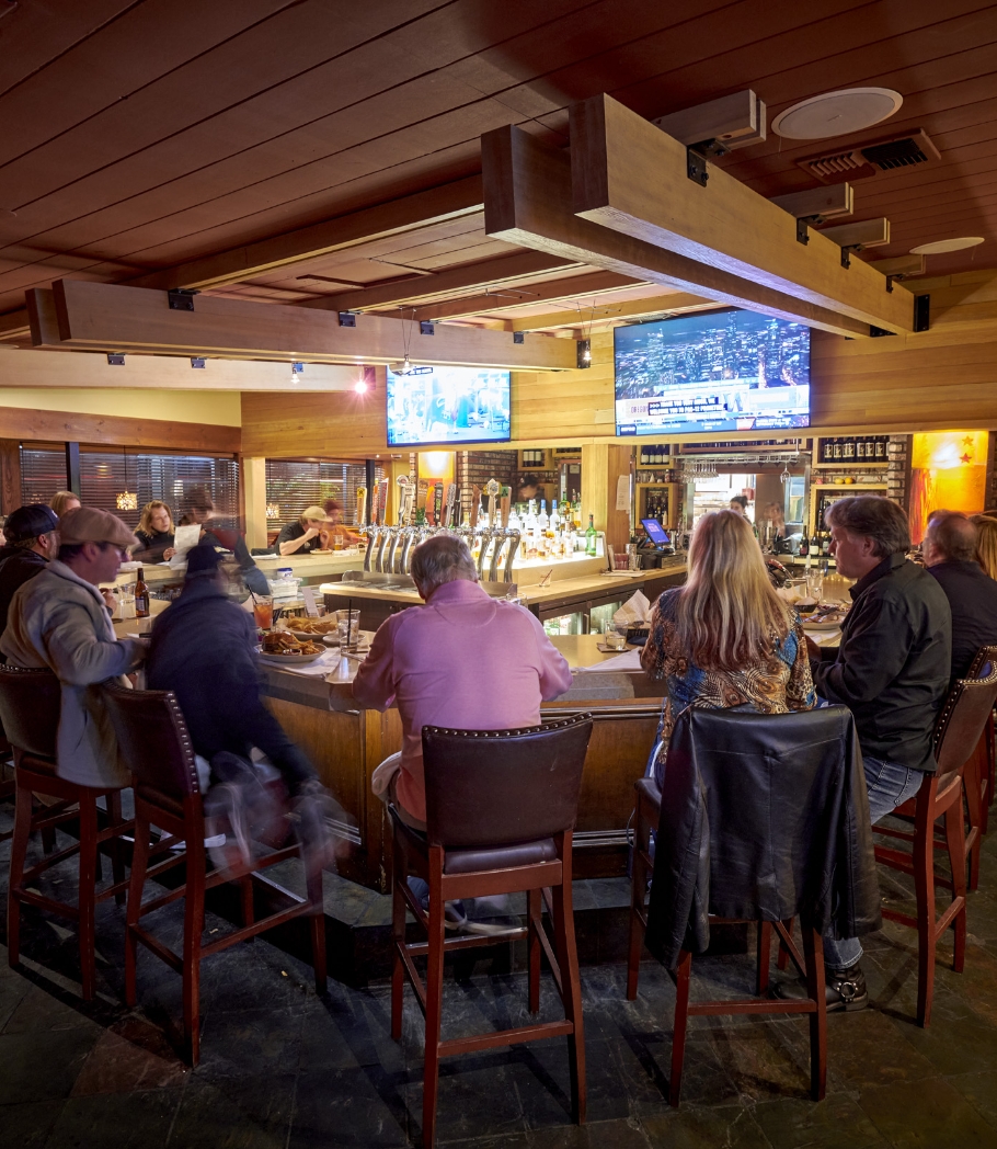Customers enjoying their time at a bar inside a restaurant with sports games broadcasting on television screens, surrounded by wood paneling and a lively atmosphere.