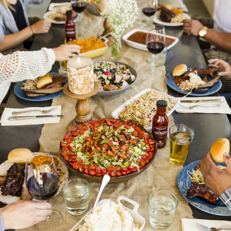 A family-style dining table filled with various dishes including a large platter of chopped salad, BBQ ribs, bowls of coleslaw, and mashed potatoes, accompanied by glasses of wine and beer. Hands are visible serving and holding plates, emphasizing a communal and cozy meal atmosphere.