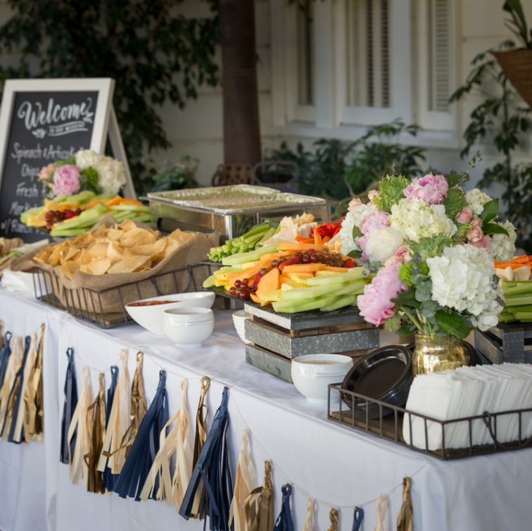 An inviting catering table at an event, featuring a colorful array of fresh cut fruit and vegetables, chips with dip, and a chalkboard sign welcoming guests to 'Spinach Artichoke Dip, Chips, Fresh Fruit.' Decorated with elegant floral arrangements and tassel table skirts adding a festive touch.