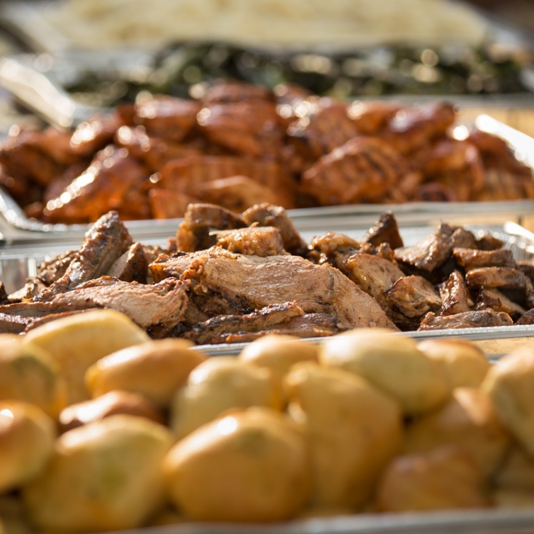 Close-up of a catering spread featuring trays of succulent grilled chicken and roasted pork, with a background of assorted cooked vegetables and golden dinner rolls, all presented enticingly under warm lighting.
