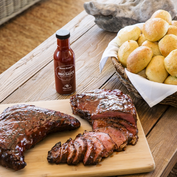 A rustic wooden table set with a cutting board displaying sliced brisket and ribs glazed with barbecue sauce, next to a bottle of Wood Ranch BBQ Sauce and a basket of freshly baked dinner rolls.