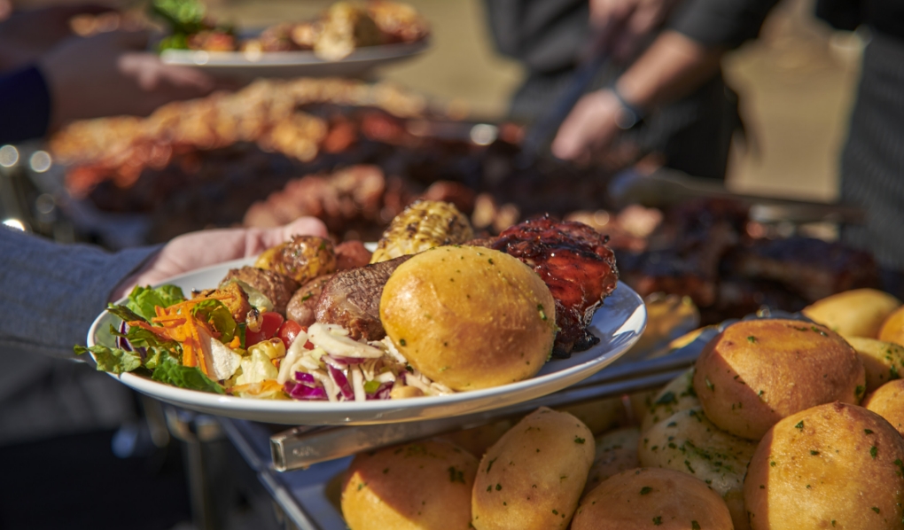 A plate held by a person featuring a variety of barbecue foods including a fresh garden salad, grilled sausage, a roll, and barbecued chicken thigh, with more food being served in the background