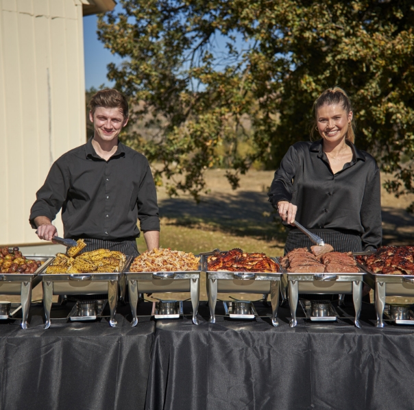 Two cheerful caterers in black attire serving a variety of grilled dishes including corn, pasta, barbecued ribs, and brisket at an outdoor buffet with a natural landscape in the background