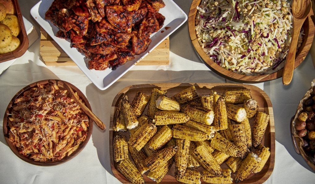 A buffet spread featuring a variety of dishes: chargrilled corn on the cob in a wooden bowl, a bowl of pasta with red sauce, barbecued chicken wings on a white platter, and a large bowl of coleslaw, all arranged on a white tablecloth under natural light.