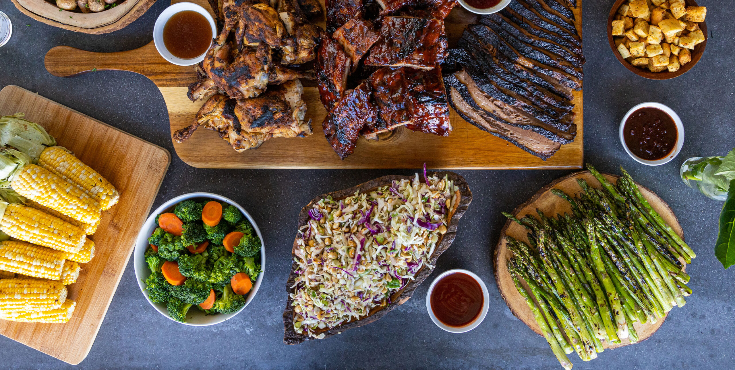 A table spread with an assortment of barbecued foods including grilled chicken, ribs, brisket, corn on the cob, coleslaw, roasted asparagus, and croutons, all beautifully presented on wooden boards and bowls, ready for a feast