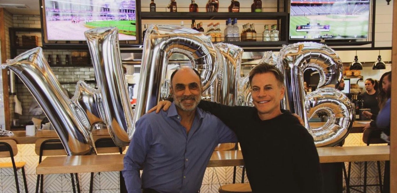 Eric Anders and Ofer Shemtov smiling and standing in front of large silver balloons spelling 'WR' inside a restaurant with a bar in the background, showing a sports game on the television screens.