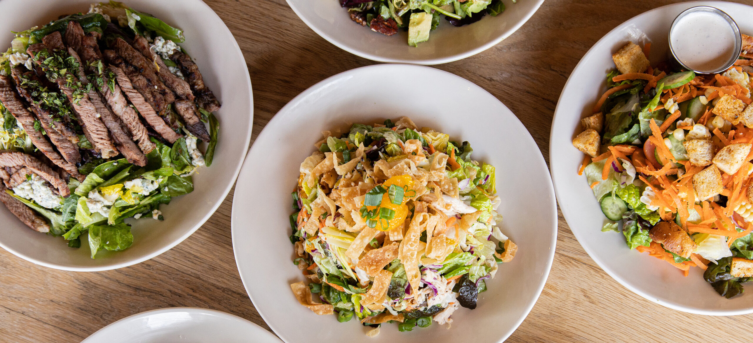 A panoramic image showcasing a variety of salads served on white plates. From left to right: a steak salad with sliced beef and greens, a colorful Asian chicken salad with mandarin oranges and crispy noodles, and a garden salad topped with croutons and shredded carrots.