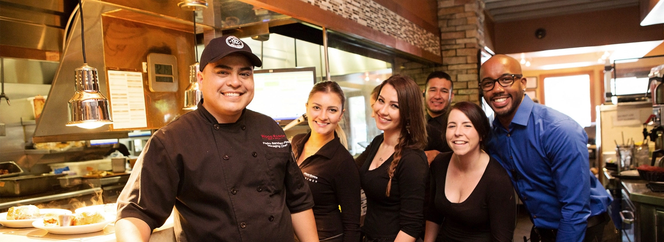 group of six restaurant staff members posing for a photo in a bustling kitchen environment. From left to right: a chef in a black uniform and cap is smiling at the camera; next to him stands a female server with her hair pulled back, smiling as well. Another server stands slightly behind them, only partly visible. In the middle, a female server with long braided hair is grinning, with another female server in front, also smiling. On the far right, a man in a blue dress shirt and glasses is leaning in, smiling with the group. In the foreground, there are plates of food ready to be served, and the background suggests a warm, inviting atmosphere with a brick wall and a softly lit dining area. The staff exude a friendly and welcoming vibe.